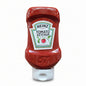 A bottle of Heinz Tomato Ketchup standing on its white cap on an orange store shelf, with warehouse shelves and other products visible in the background.